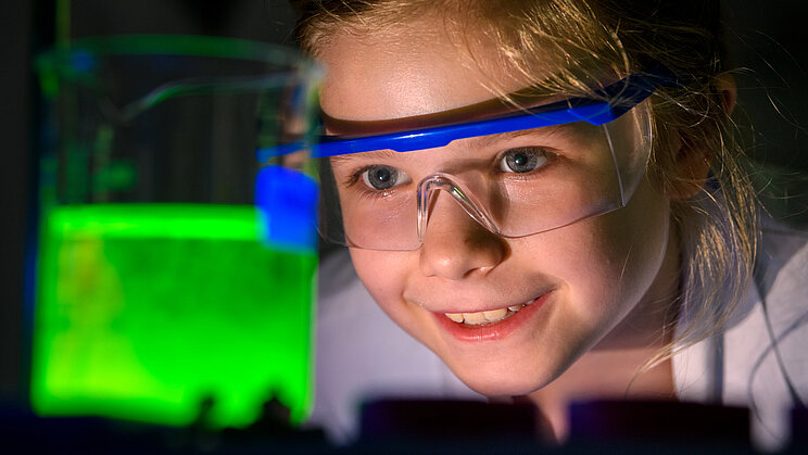 Es ist ein Mädchen abgebildet, die ein Reagenzglas mit einer grünen, leuchtenden Flüssigkeit beobachtet